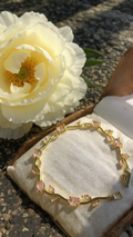 A gold bracelet with pink marquise-cut gemstones displayed on a cream jewelry cushion inside an open wooden box, with a large white flower beside it on a stone surface.
