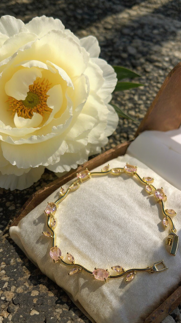 A gold bracelet with pink marquise-cut gemstones displayed on a cream jewelry cushion inside an open wooden box, with a large white flower beside it on a stone surface.