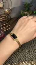 A gold-tone mesh bracelet watch with a rectangular black dial and a gold bezel, shown worn on a person's wrist against a home interior background.