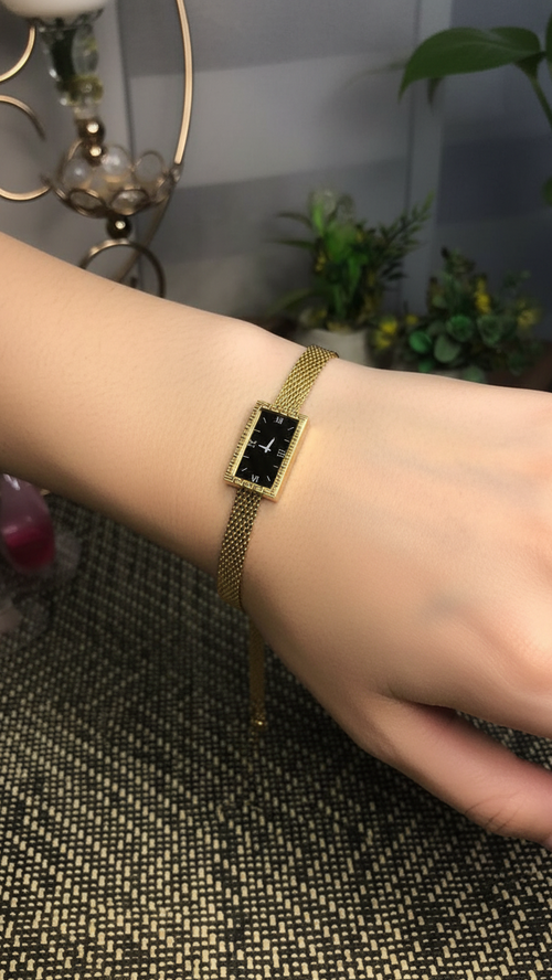 A gold-tone rectangular watch with a black dial and a gold mesh band, shown worn on a person's wrist against a softly blurred indoor background with plants and home decor.