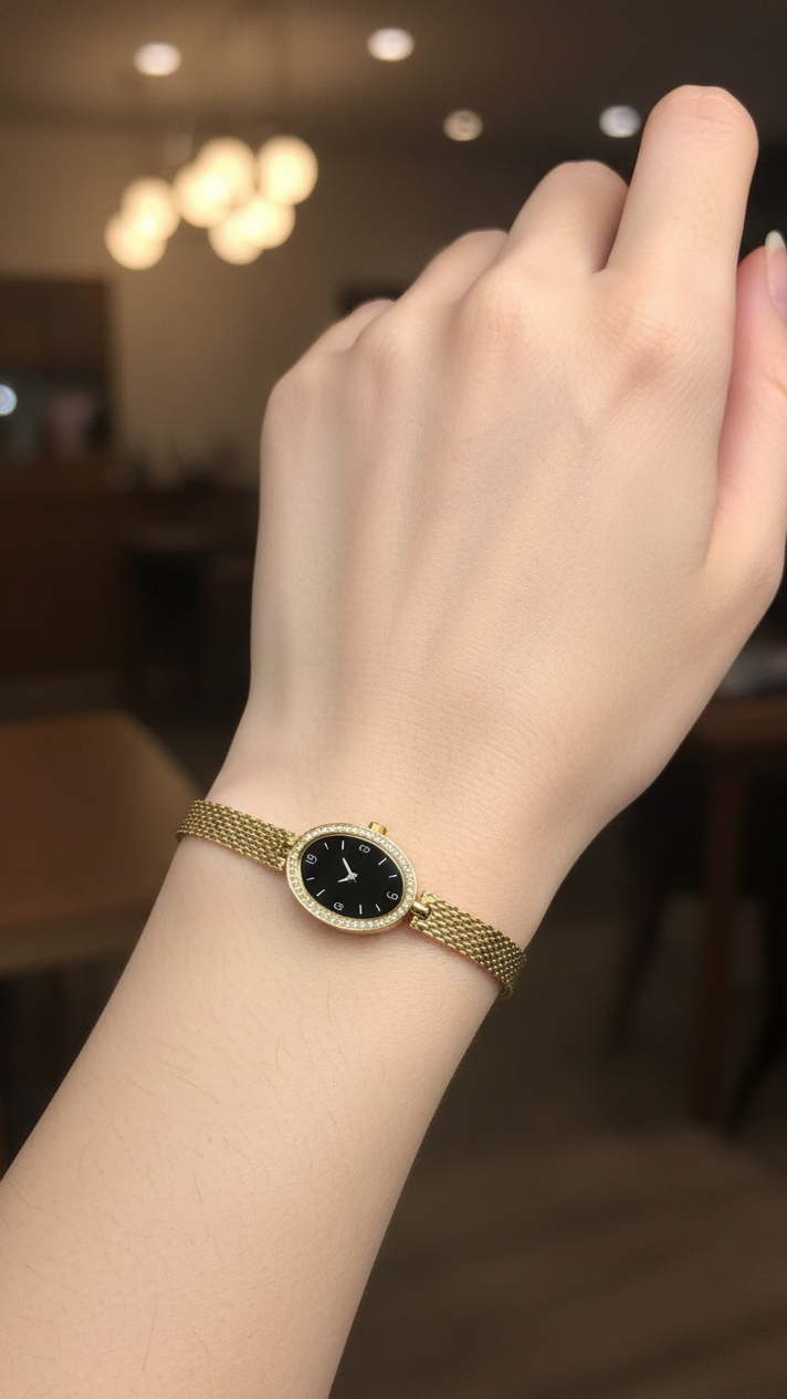A gold mesh bracelet watch with a black dial and a bezel set with small clear stones, shown worn on a person's wrist in a softly blurred indoor setting.