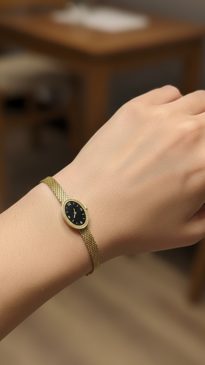 A gold mesh bracelet watch with a black dial and gold bezel, shown worn on a person's wrist against a blurred indoor background.