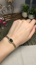 A gold mesh bracelet watch with a round black dial and gold bezel, shown worn on a person's wrist. The background features a wooden table, decorative plants, and home decor items.