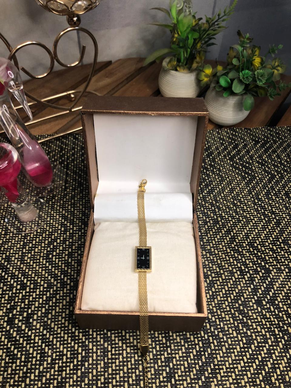 A gold-tone watch with a rectangular black face is displayed inside an open brown presentation box on a white cushion, set on a patterned surface with decorative plants and glassware in the background.