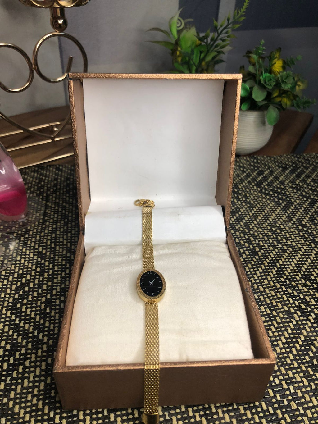 A gold-tone mesh bracelet watch with a round black dial, displayed inside an open brown presentation box with a white cushion.
