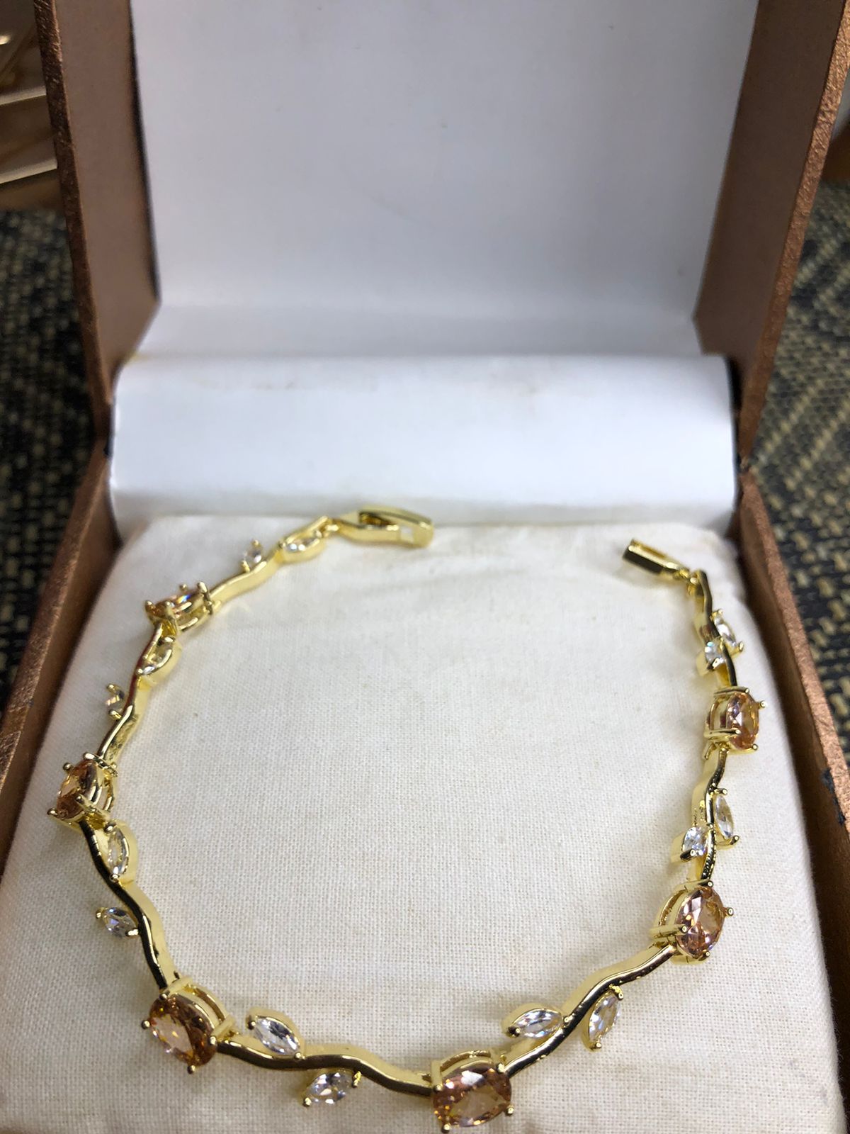 A gold bracelet with clear and peach-colored crystals, displayed inside an open brown jewelry box with a cream-colored lining.